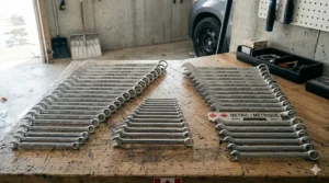 A comprehensive photorealistic illustration fanning out over fifty SAE and metric combination wrenches from a larger combo set, organized on a wooden bench in a Canadian garage.